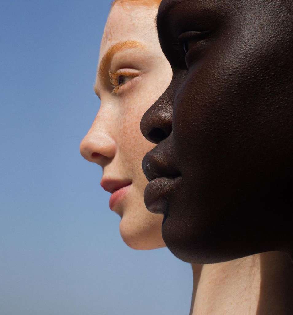 Close-up of two women with contrasting skin tones standing side-by-side against a clear blue sky. Portrait showcases diversity and beauty.