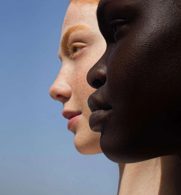 Close-up of two women with contrasting skin tones standing side-by-side against a clear blue sky. Portrait showcases diversity and beauty.