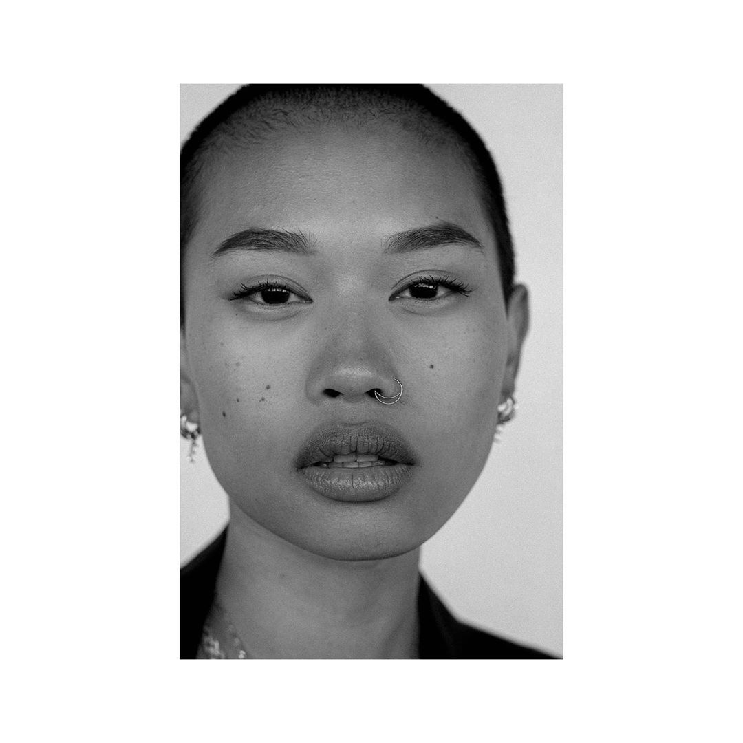 Close-up portrait of a young adult woman with shaved head, a nose ring, wearing a black jacket against a white backdrop in black and white.