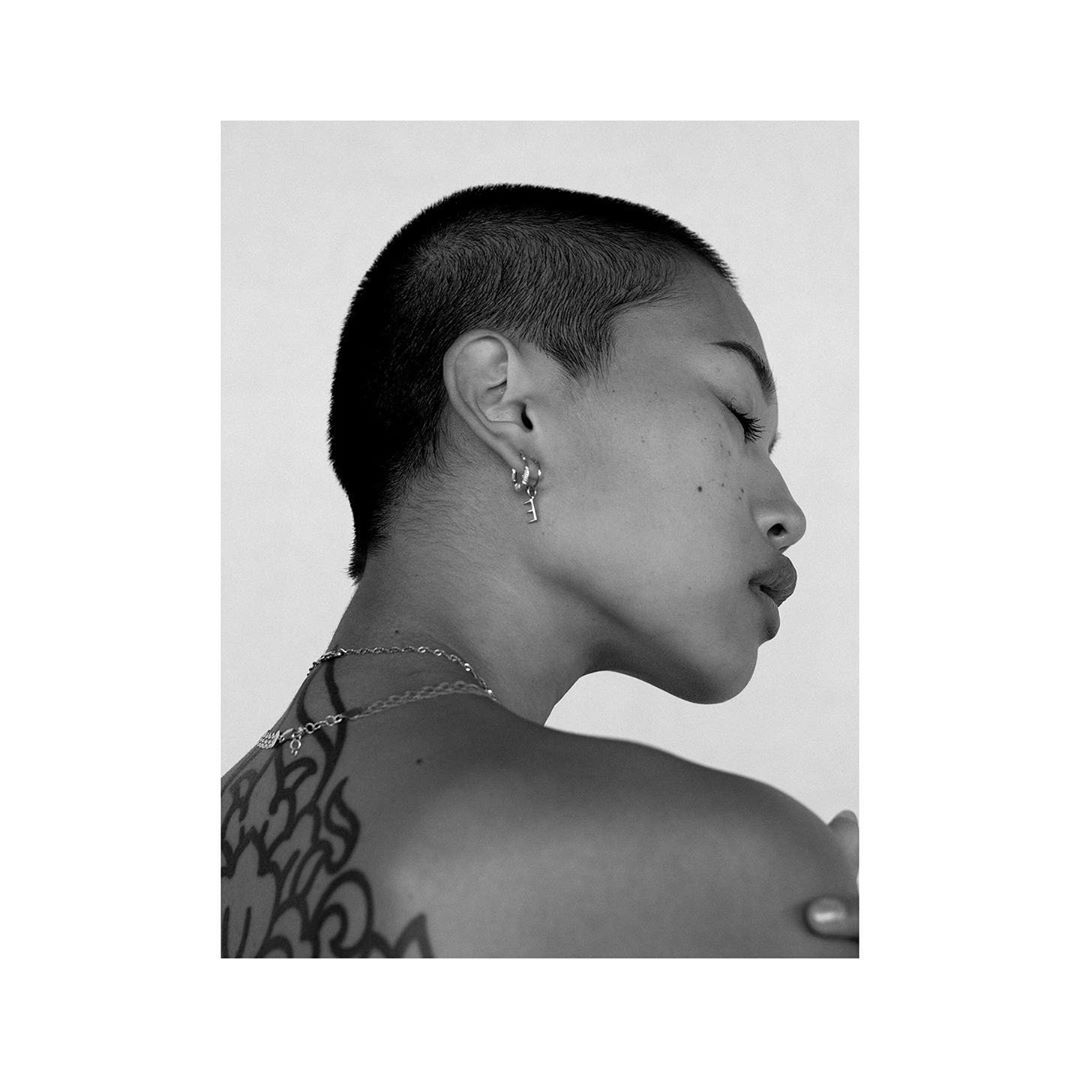 Striking black and white portrait of a woman with short hair, featuring her tattoo, silver jewelry, and minimalistic fashion against a plain background.