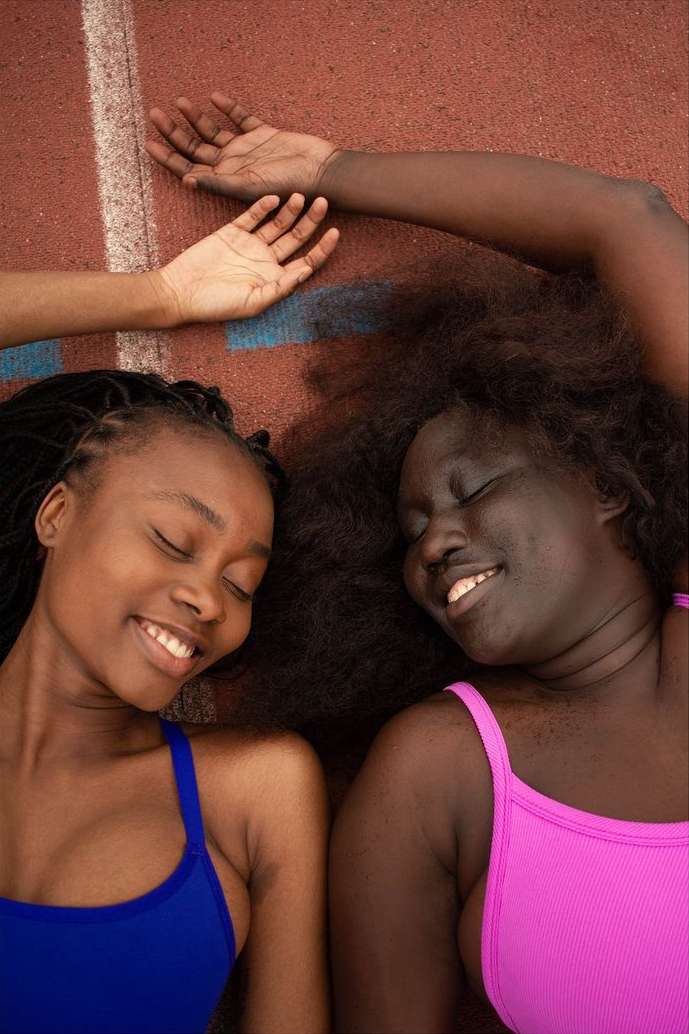 Two smiling African American women in brightly colored tops lie side by side on an orange running track with a single white stripe.