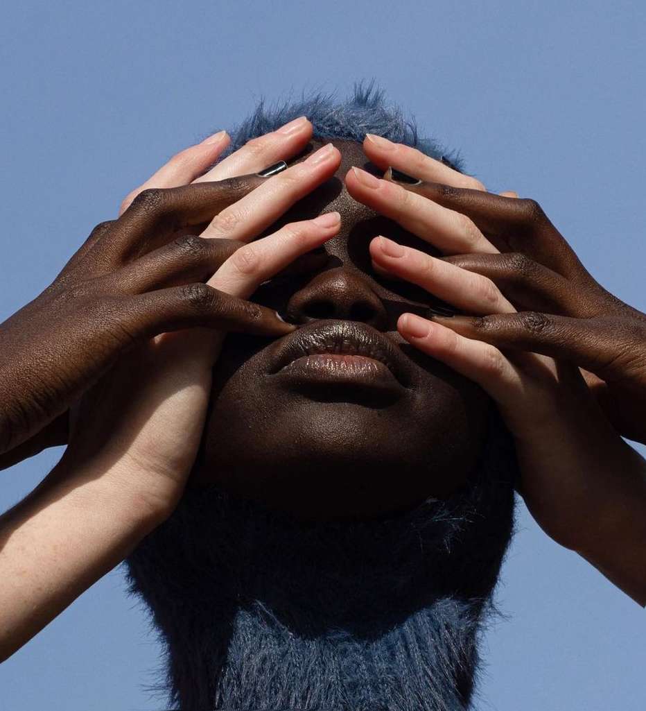 A person with dark skin and blue hair is covering their face with hands of lighter skin tones against a blue sky background.