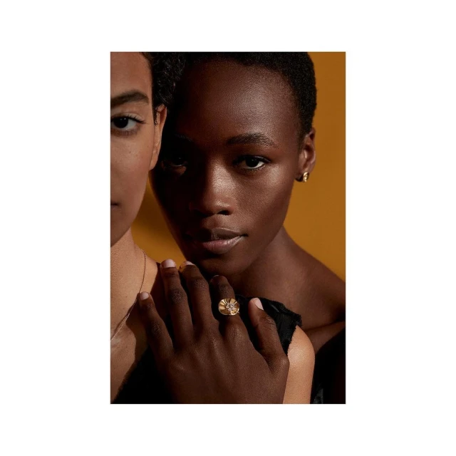 Close-up beauty shot of two adult women with warm skin tones and gold ring, set against a warm yellow backdrop in a photo studio.