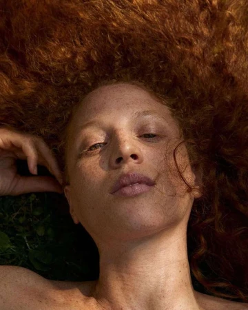 Close-up portrait of a woman with freckles and voluminous red curly hair captured from an overhead angle.