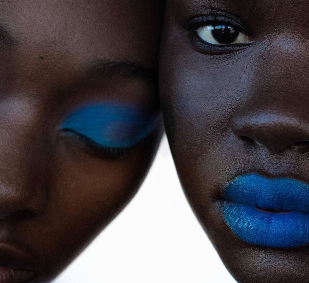 Close-up of two women with dark skin. One woman has bright blue eyeshadow and the other has matching blue lipstick.