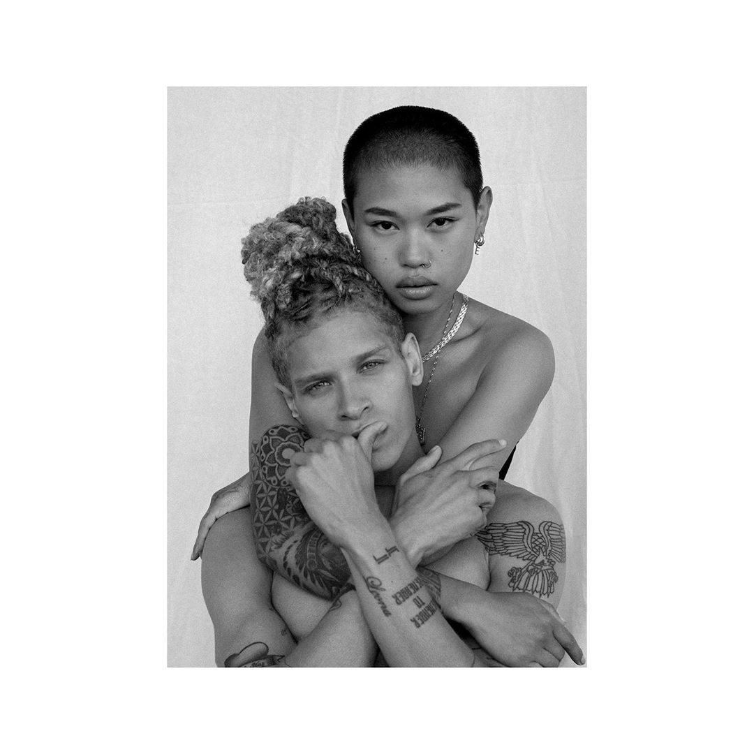 Black and white studio portrait of a bare-chested male with arm tattoos being embraced by a buzz-cut female wearing a chain necklace.