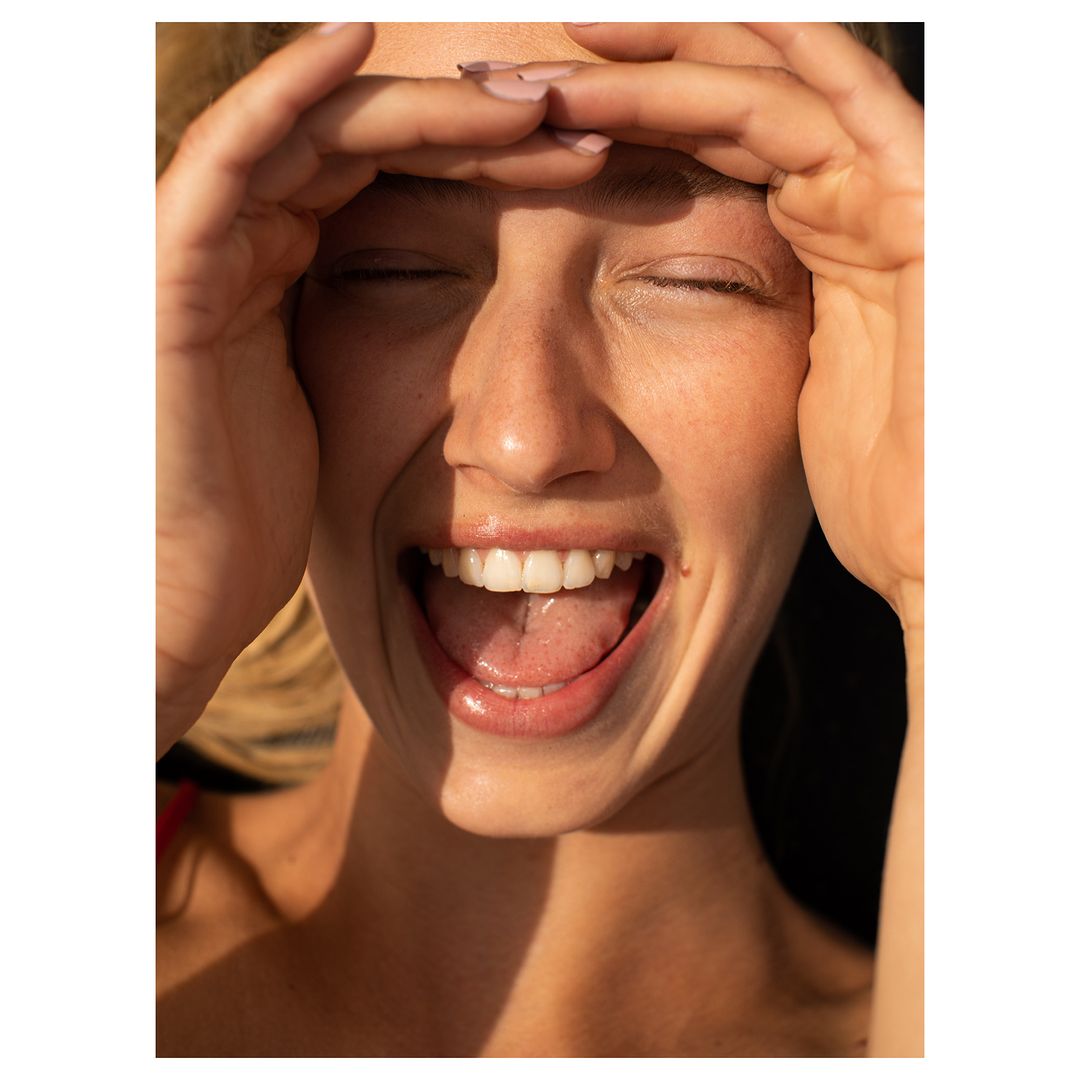 Close-up portrait of a smiling woman with her tongue out, hands framing her face, eyes closed, bathed in warm, golden sunlight.