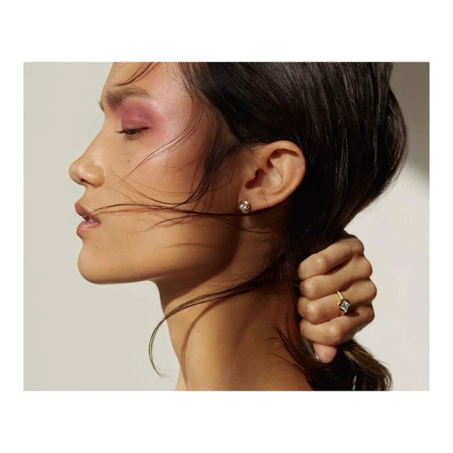 Profile of an adult female wearing jewelry, featuring a ring and earrings, with soft pink eye makeup and a neutral background.