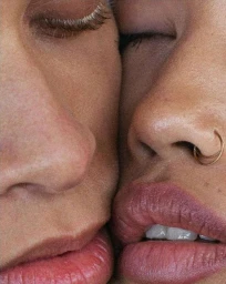 Close-up macro portrait of two people pressing their faces together with visible skin texture, lips, and a gold nose ring.