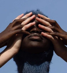 Close-up portrait of a dark-skinned person with blue hair surrounded by multiple hands covering their face against a blue sky.
