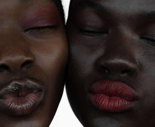 Close-up portrait of two Black women with closed eyes touching cheeks, featuring deep red lipstick and burgundy eyeshadow.