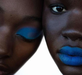 Close-up portrait of two Black women with vibrant blue eyeshadow and bright blue lipstick against a clean white background.