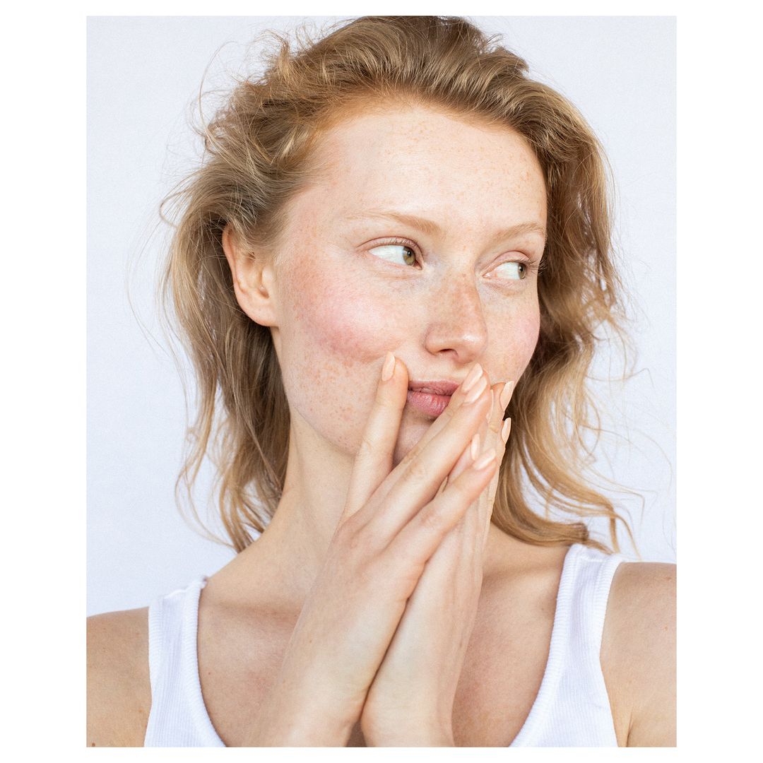 Close-up portrait of a woman with red hair and freckles holding her hands near her face against a white background