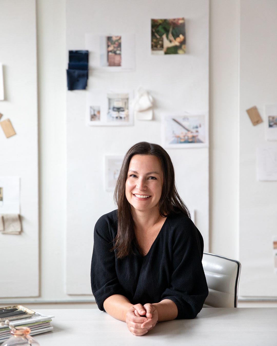 Smiling woman with dark hair at a white desk in an office. Behind her are pinned images and color swatches on white boards.