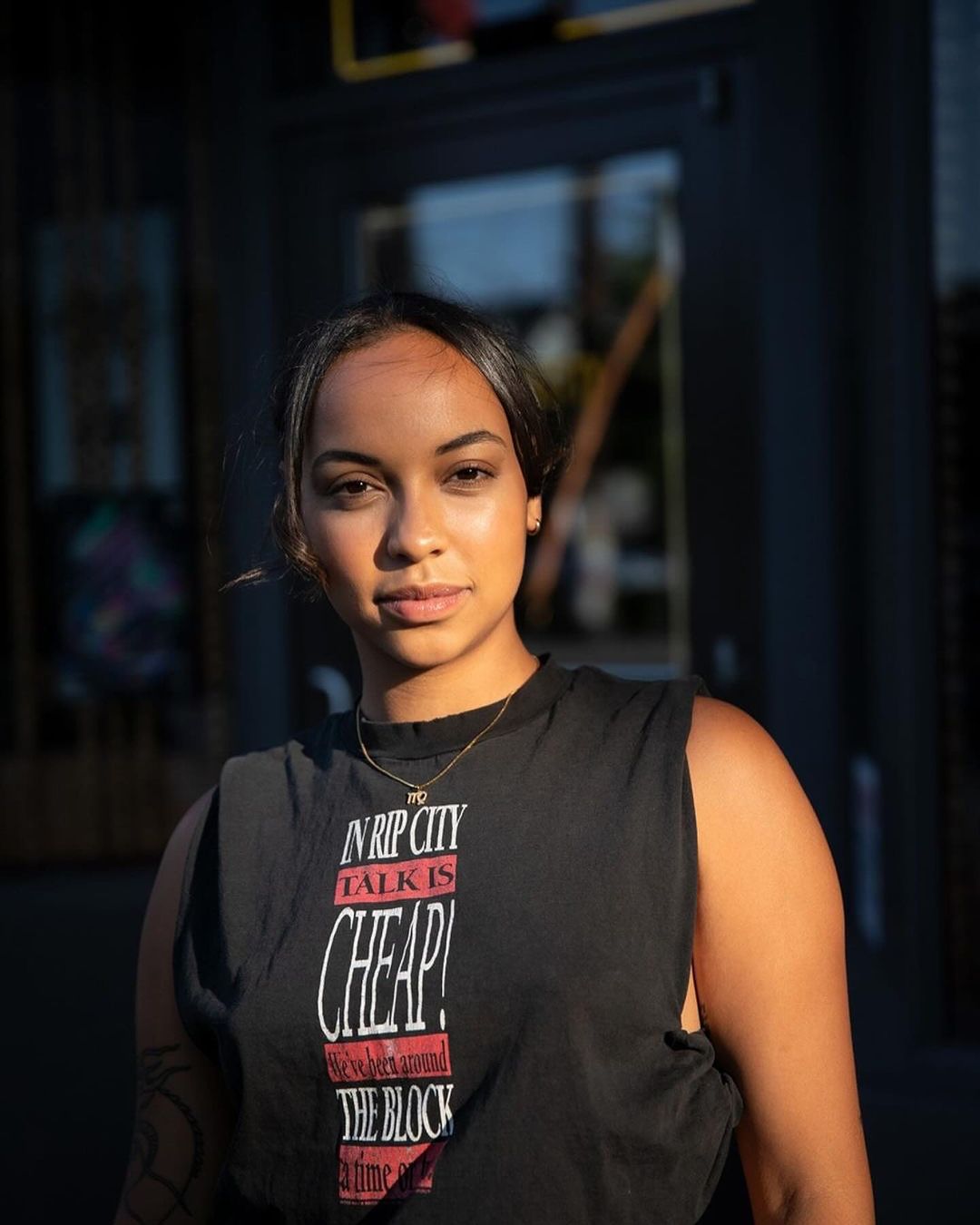 Close-up shot of a young woman wearing a black t-shirt with graphic text standing outdoors in front of a dark building.