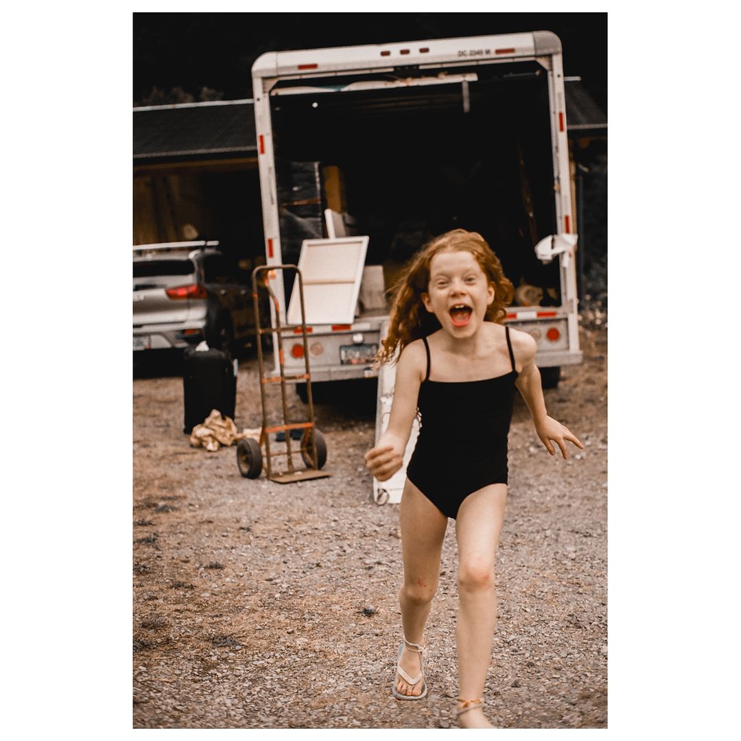A young girl with red hair runs outside wearing a black bathing suit with a utility dolly and white truck in the background.