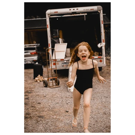 A young girl with red hair runs outside wearing a black bathing suit with a utility dolly and white truck in the background.