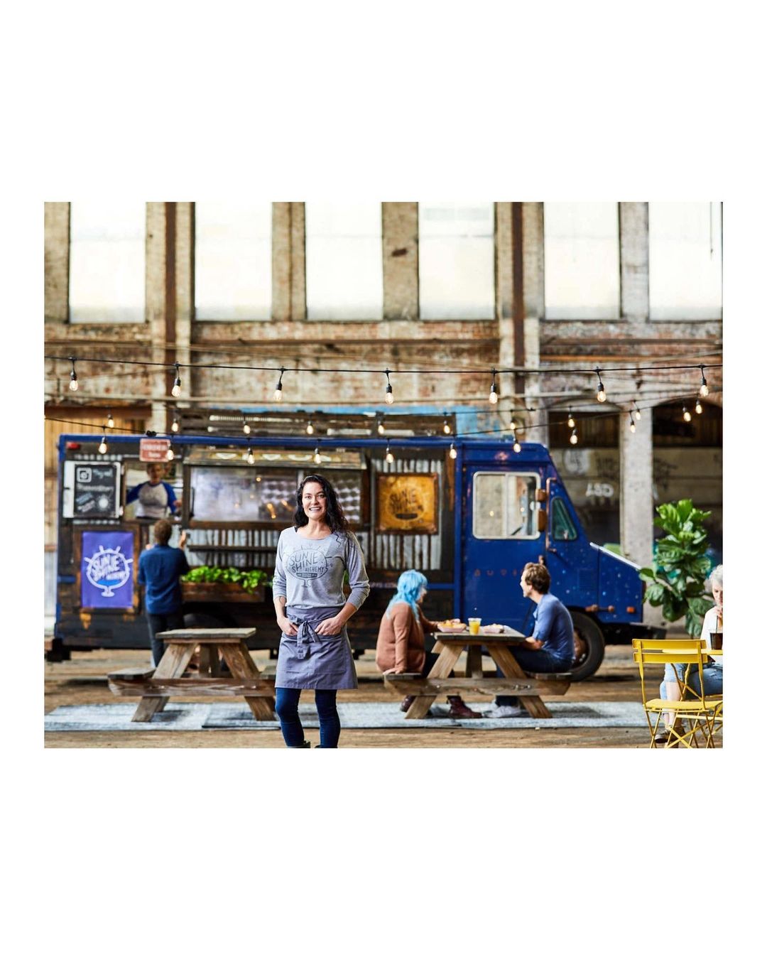 Woman in apron stands in front of food truck with customers at picnic tables, illuminated by string lights in urban setting.