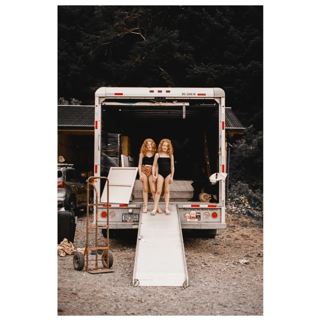 Two young girls in bathing suits stand on the ramp of a moving truck with a forest backdrop, creating a candid, summery scene.