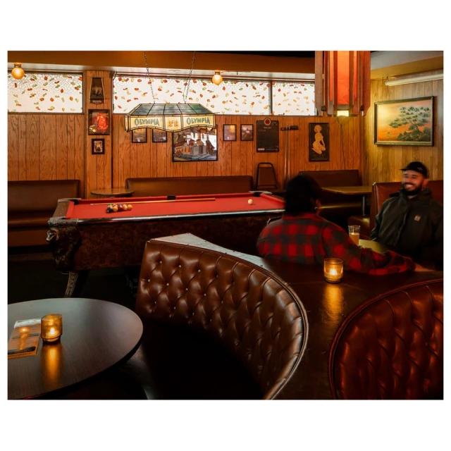 Interior shot of a vintage bar with a red pool table, brown leather booths, and two adult men sitting at a table with dim lighting.
