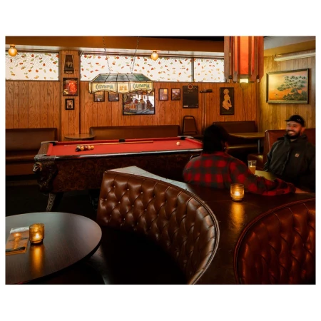 Interior shot of a vintage bar with a red pool table, brown leather booths, and two adult men sitting at a table with dim lighting.