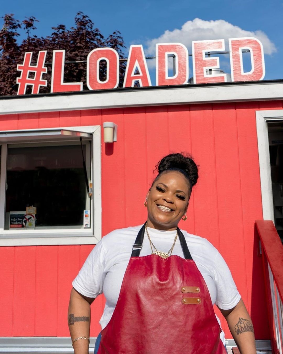 Smiling woman in a red apron stands in front of a red food truck with '#LOADED' on top, under a partly cloudy blue sky.