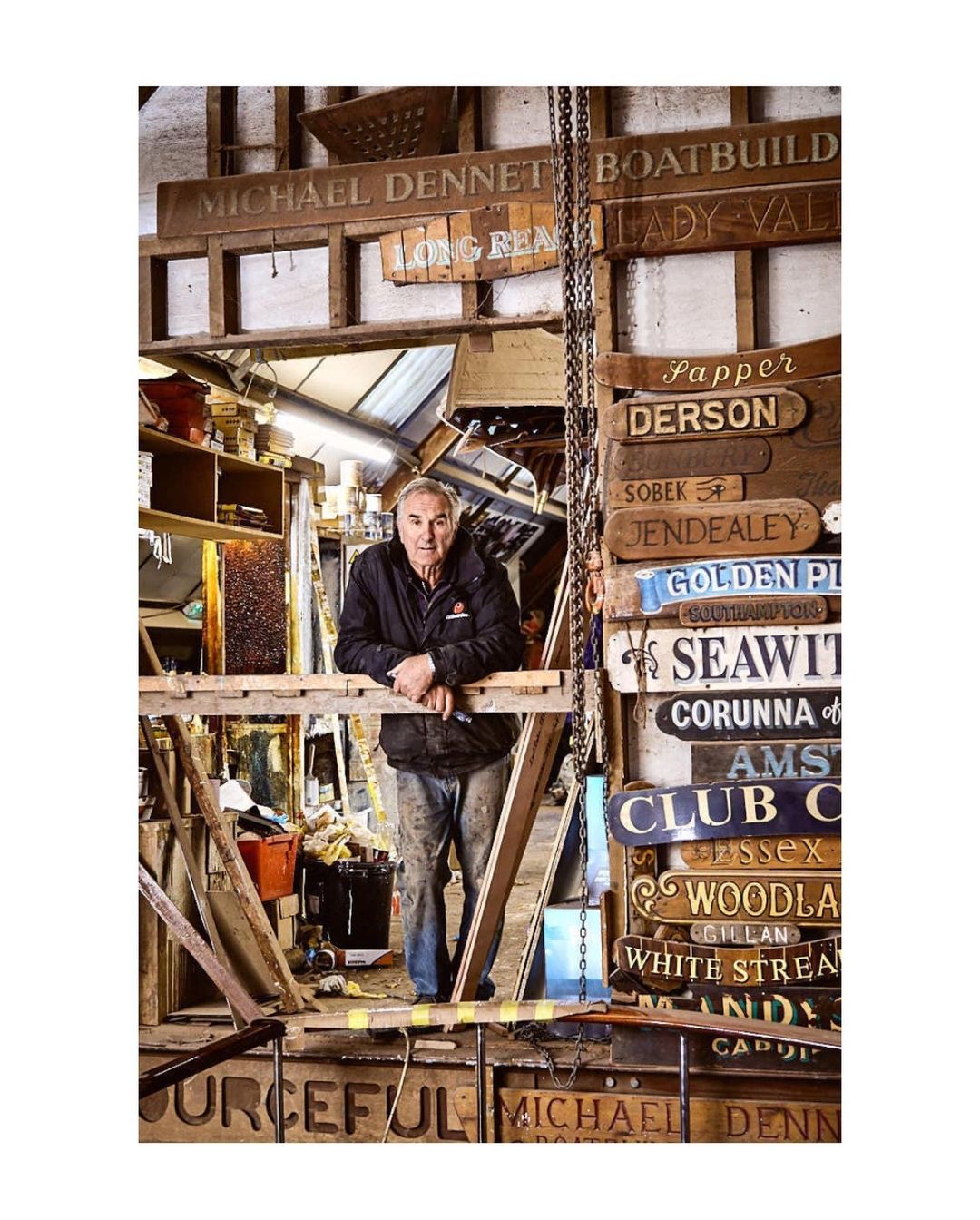 An adult male stands in a woodworking shop surrounded by hand-carved, wooden signs, exuding a rustic, handcrafted, and personalized ambiance.