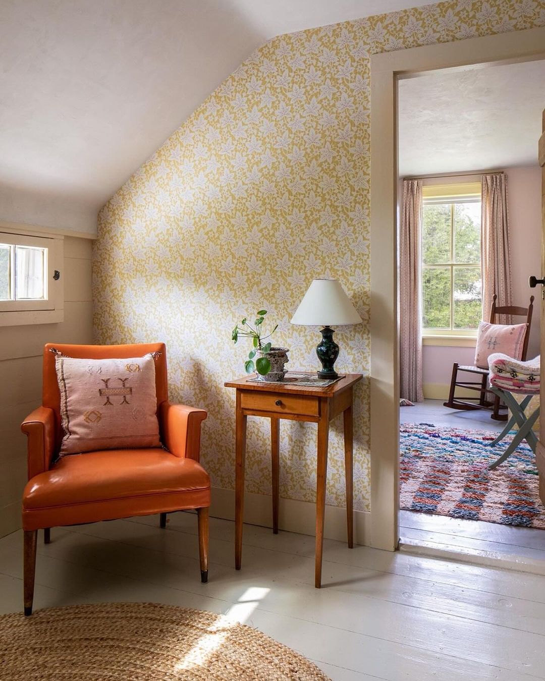 Cozy interior with an orange armchair and yellow floral wallpaper, lit by a table lamp next to a doorway leading to another room.