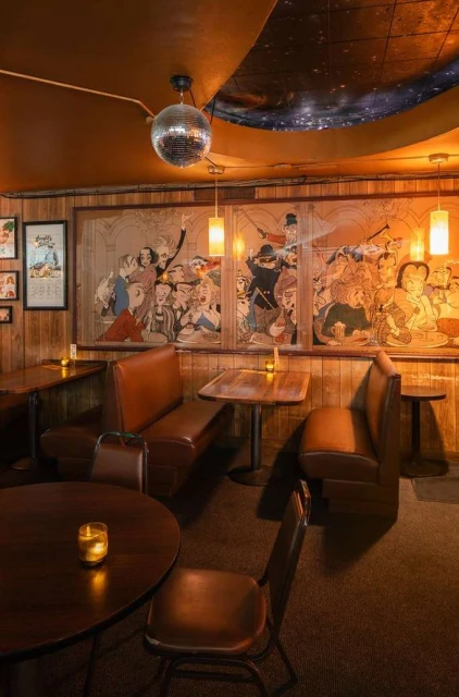 Retro interior of a lounge or bar featuring booths and tables, a mirrored disco ball, and artwork, creating a vintage, ambient setting.