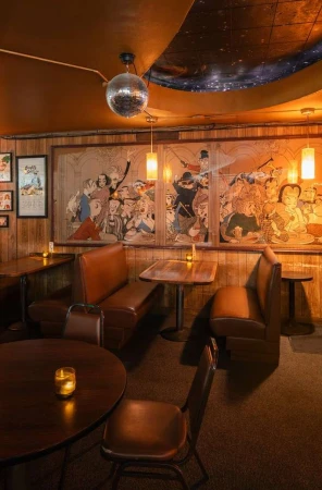 Retro interior of a lounge or bar featuring booths and tables, a mirrored disco ball, and artwork, creating a vintage, ambient setting.