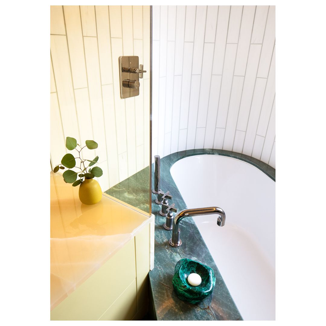 Modern bathroom features a white bathtub with green marble accents, chrome fixtures, and a yellow vase with green leaves on a vanity.
