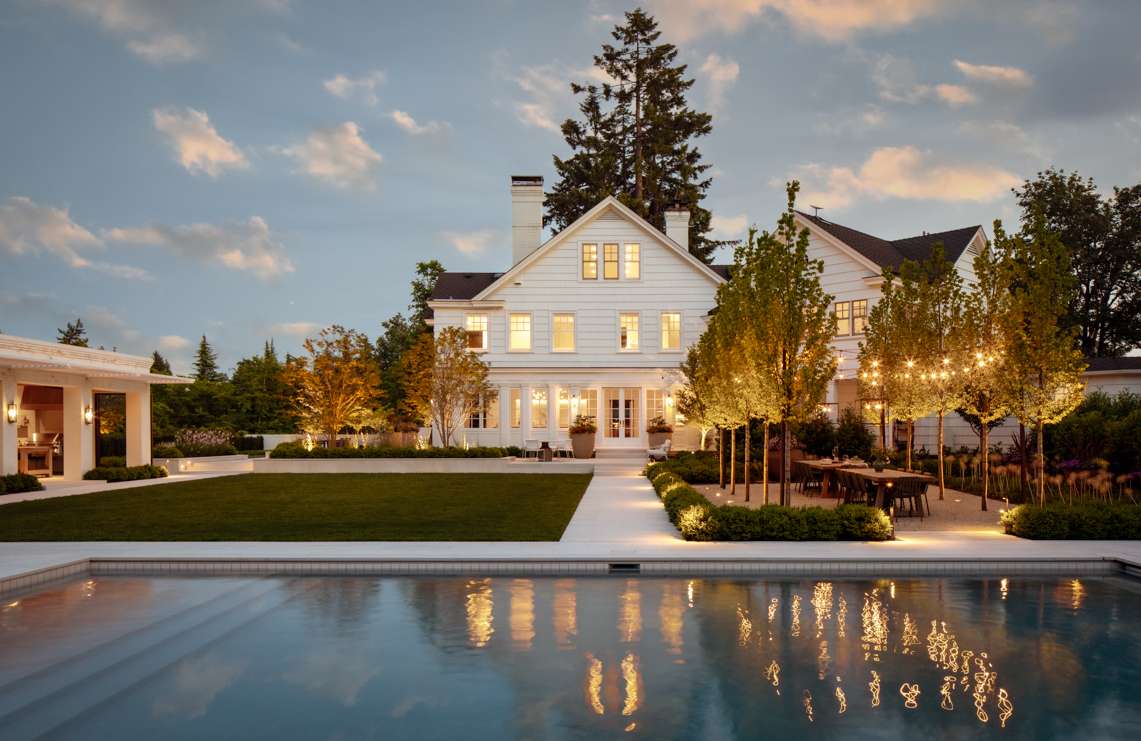 Exterior view of a modern white farmhouse with a pool, landscaped lawn, trees, and outdoor dining area at dusk, showcasing home design.
