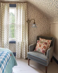 A grey armchair with patterned throw pillows in a bedroom with blue floral wallpaper and white curtains.