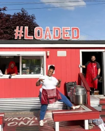 Three smiling food workers pose outside a bright red loaded food shack with a prominent sign under a clear sky.