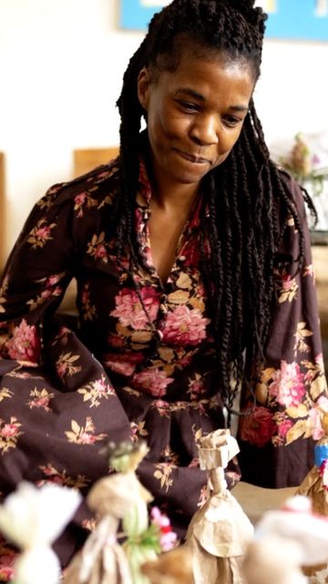 Adult female with long dark braids is gazing downward while seated in front of several wrapped-paper figures inside a brightly lit room.