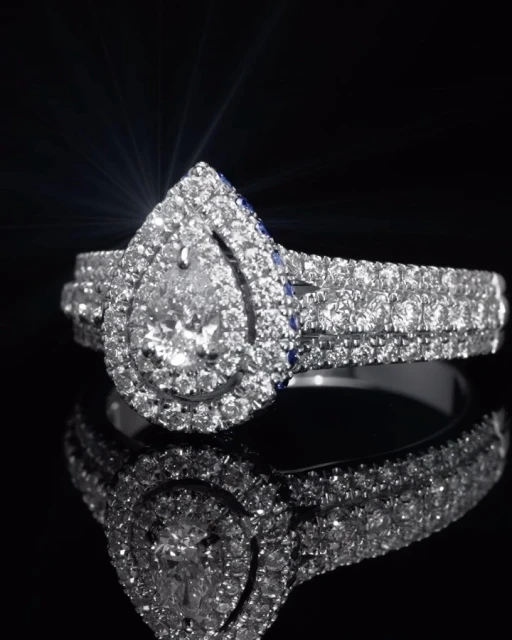 Sparkling pear-shaped diamond ring with a halo setting, set against a dark reflective surface, emphasizing its brilliance and design.