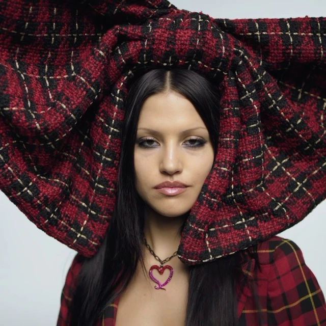 Headshot of a woman with long black hair wearing a red and black plaid blazer and a large matching bow, accessorized with a heart necklace.