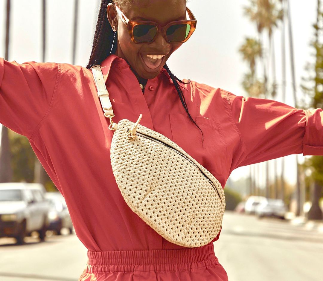 A cheerful adult woman in a red jumpsuit and sunglasses stands outside with a cream-colored woven belt bag, surrounded by palm trees.
