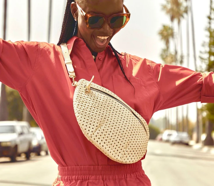 A cheerful adult woman in a red jumpsuit and sunglasses stands outside with a cream-colored woven belt bag, surrounded by palm trees.