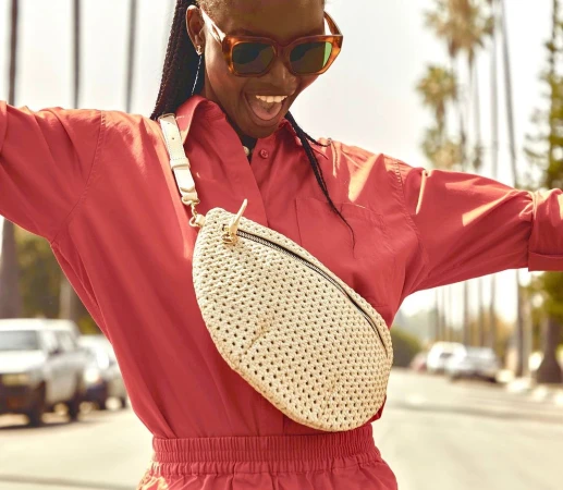 A cheerful adult woman in a red jumpsuit and sunglasses stands outside with a cream-colored woven belt bag, surrounded by palm trees.