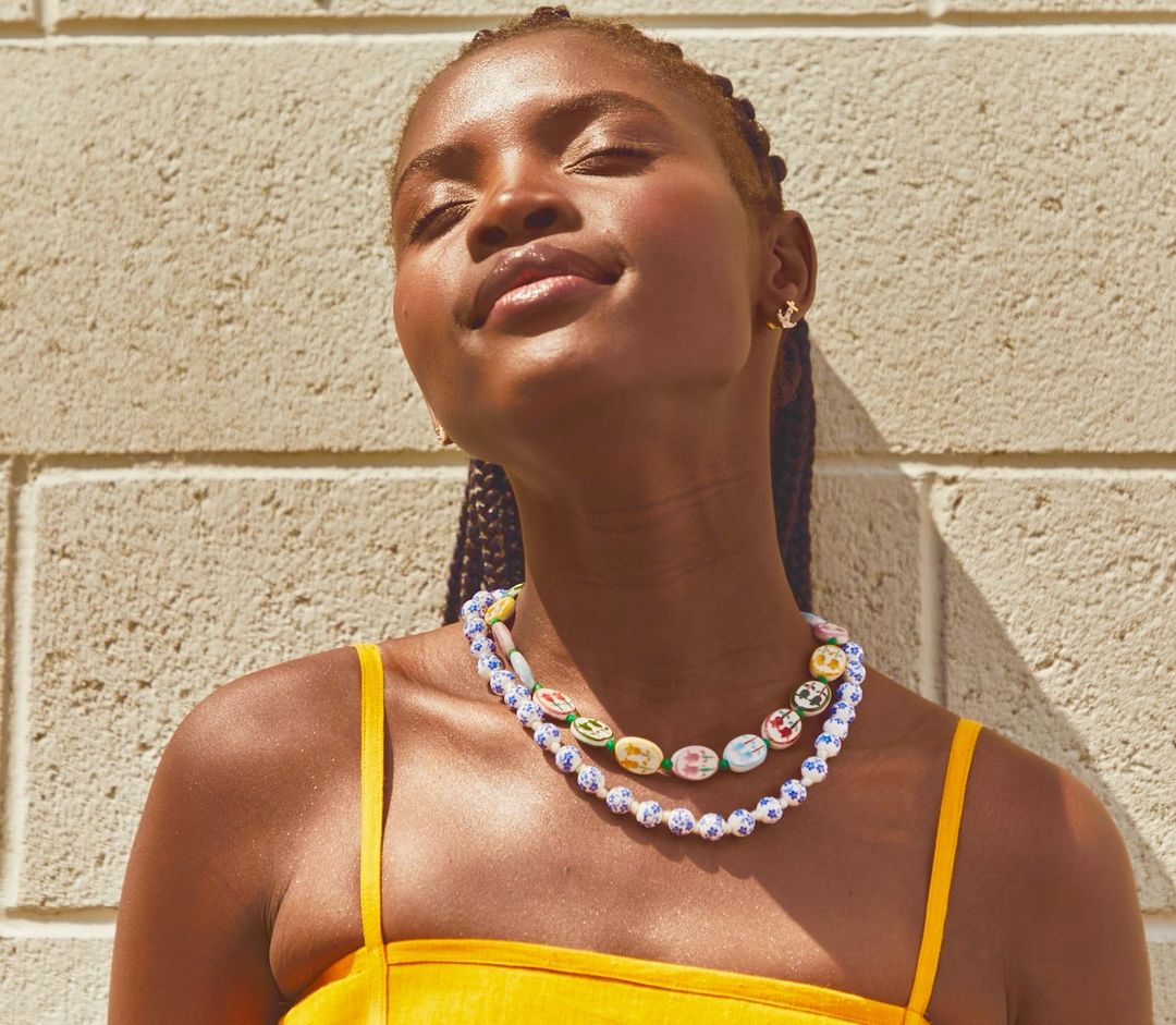 Portrait of an African American woman with braided hair in a yellow dress against a textured wall, wearing colorful beaded necklaces.
