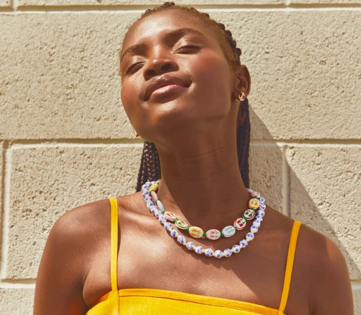 Portrait of an African American woman with braided hair in a yellow dress against a textured wall, wearing colorful beaded necklaces.