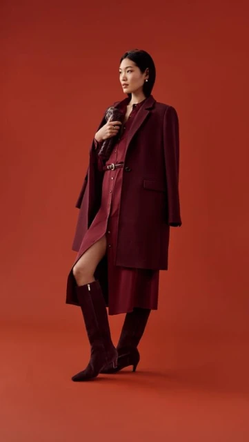 A stylish Asian woman with short dark hair poses in a burgundy dress, coat, and boots against a matching backdrop in a full shot.