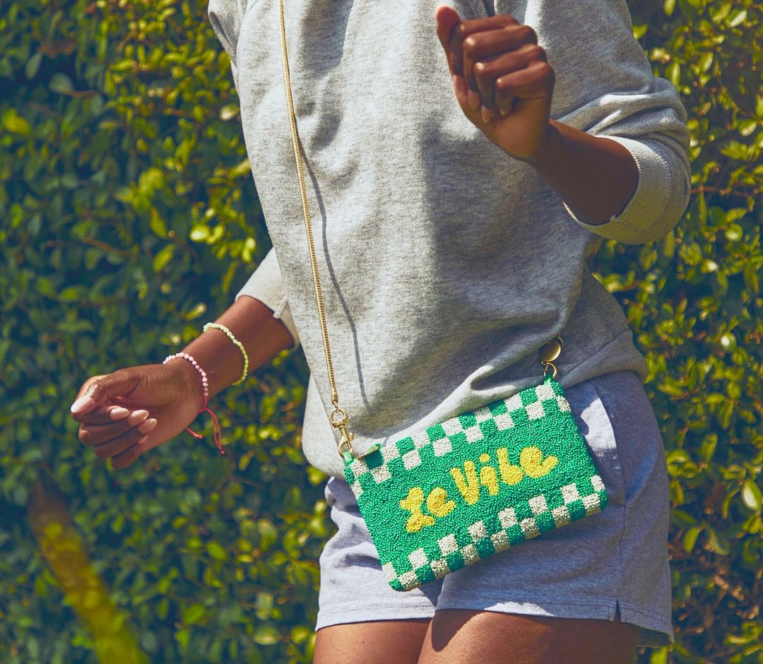 A fashionable green checkerboard purse with 'Le Vibe' text, worn by a person in a grey sweatshirt outdoors against a leafy backdrop.