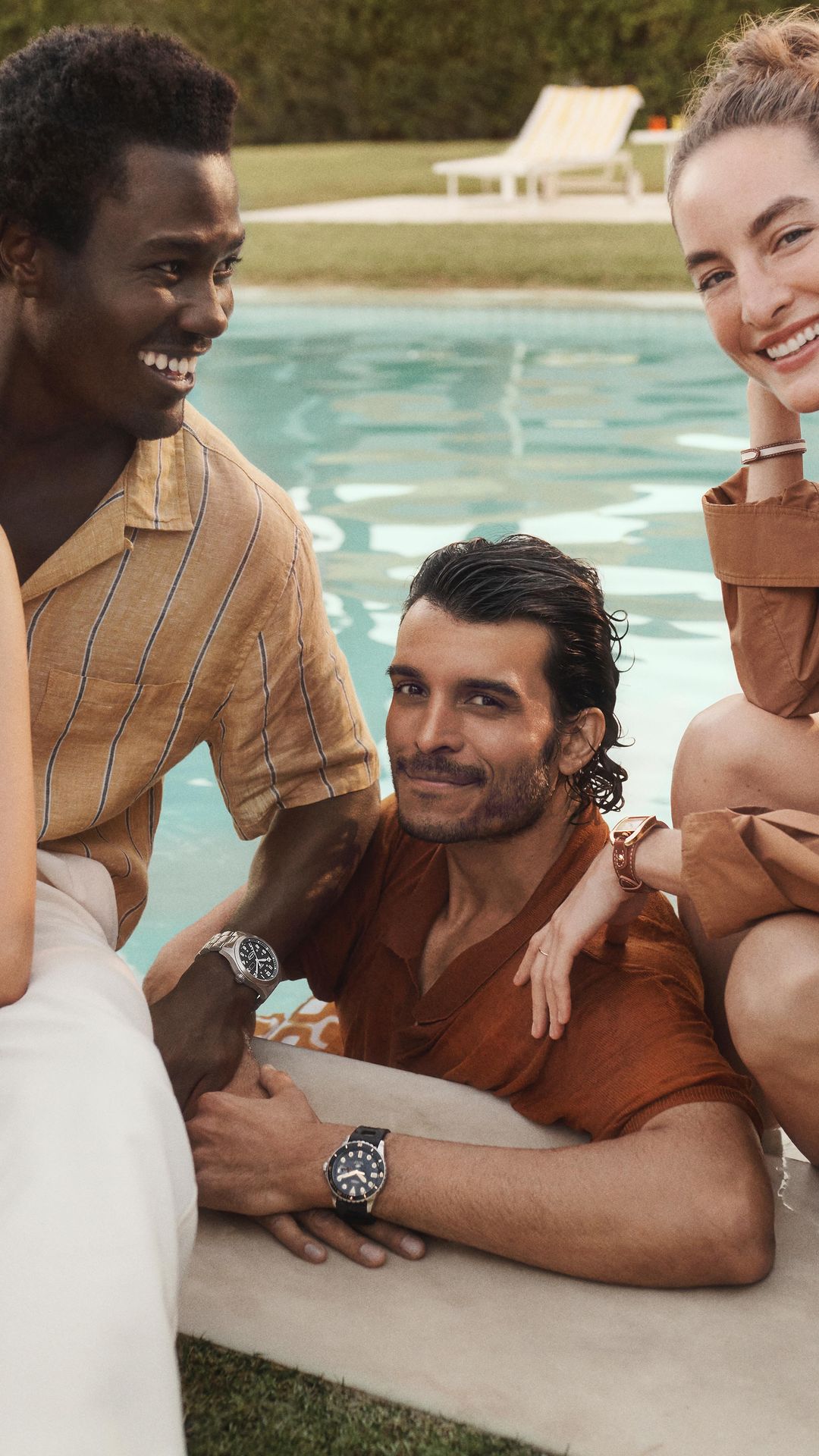 Group of smiling people gathered outdoors by a pool. Includes adult males and females wearing fashionable clothing and wristwatches.