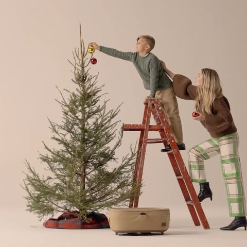 A young boy on a ladder decorating a Christmas tree with the assistance of a woman, set against a clean studio backdrop.