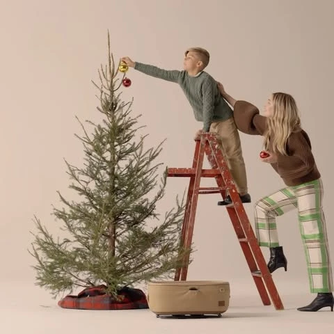 A young boy on a ladder decorating a Christmas tree with the assistance of a woman, set against a clean studio backdrop.