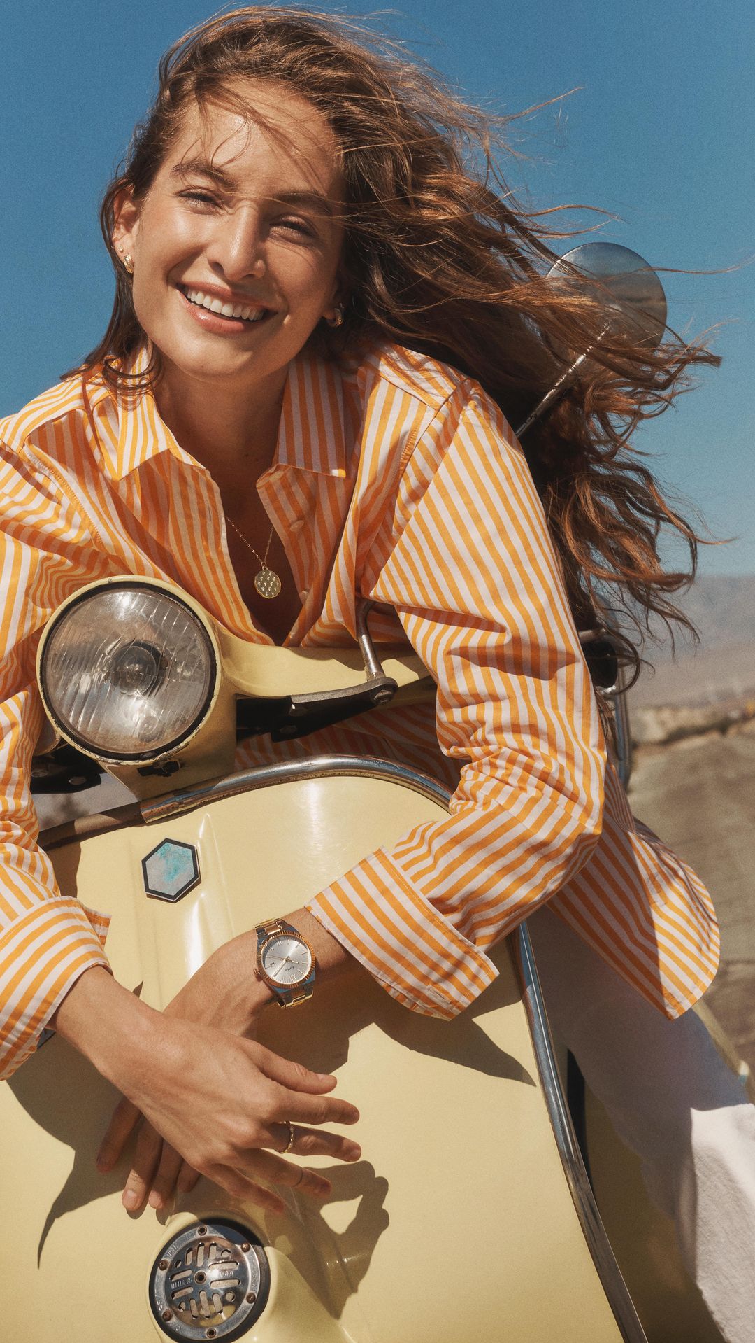 Smiling woman poses with a pale yellow scooter on a clear day; she wears an orange striped shirt and stylish wristwatch.