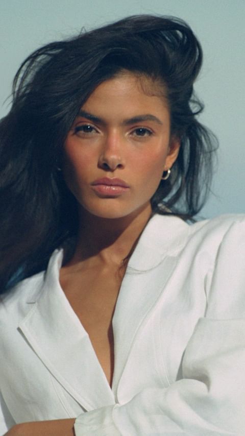 Brunette woman in white blazer poses against a light blue background in a beauty shot with soft lighting, and a shallow depth of field.