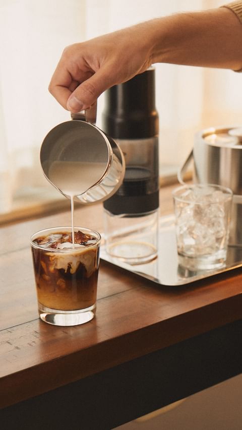 Cream is poured into a glass of iced coffee on a wood table with a stainless-steel thermos and glasses on a silver tray nearby.